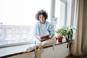 Young man using tablet PC on window sill at home - NLAF00234