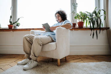 Man sitting on armchair and using tablet PC at home - NLAF00235