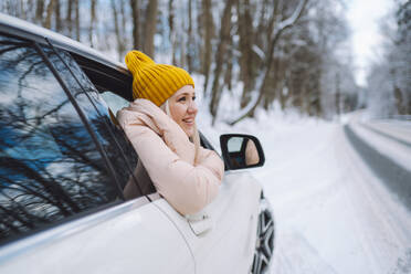 Frau mit Strickmütze, die sich im Winterwald aus dem Autofenster lehnt - OLRF00120