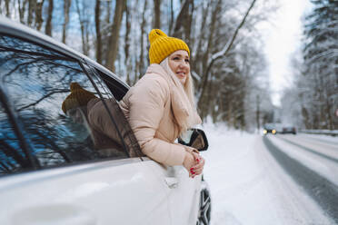Happy woman leaning out of car window at roadside in winter forest - OLRF00122