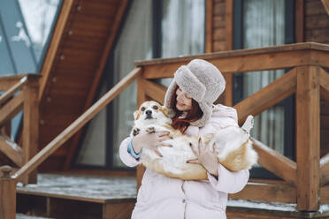 Smiling young woman holding Corgi dog in front of log cabin - OLRF00128