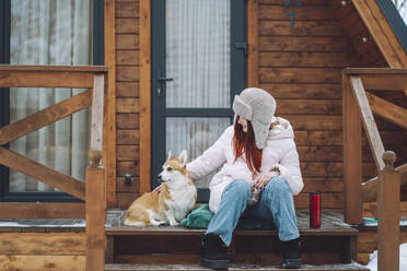 Glückliche Frau mit Corgi-Hund auf der Veranda einer Blockhütte - OLRF00129