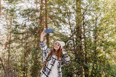 Happy woman taking selfie through smart phone standing in front of tree - EBBF08295