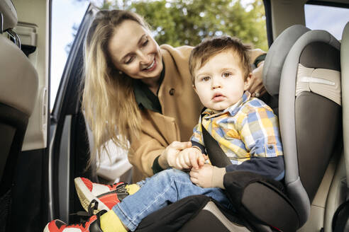 Lächelnde Mutter, die den Sicherheitsgurt für ihren im Auto sitzenden Sohn einstellt - NJAF00742
