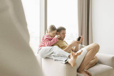 Mädchen sitzt in der Nähe des Vaters und benutzt ein Smartphone zu Hause - SEAF02184
