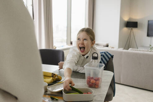 Playful girl stealing food from lunch box on table at home - SEAF02195