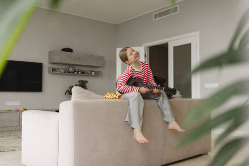Glückliches Mädchen sitzt auf dem Sofa und spielt Spielzeuggitarre zu Hause - SEAF02242