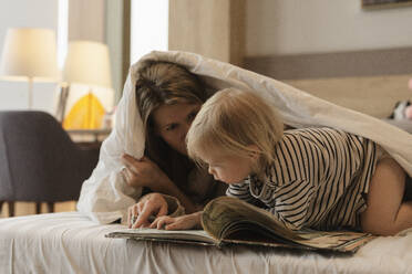 Mother reading book to daughter under blanket in bed at home - SEAF02244