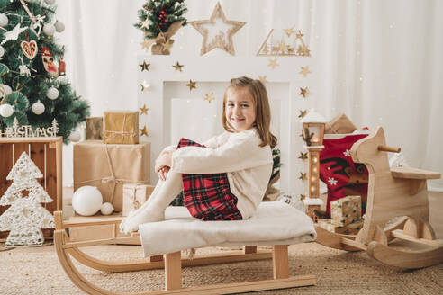 Lächelndes Mädchen sitzt auf einem Schlitten in der Nähe von Weihnachtsschmuck zu Hause - EBBF08306