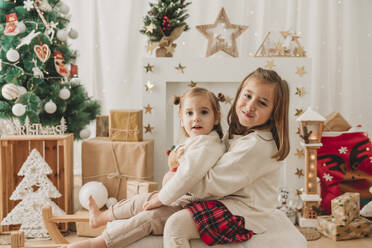 Glückliche Schwestern sitzen auf einem Schlitten in der Nähe der Weihnachtsdekoration zu Hause - EBBF08313