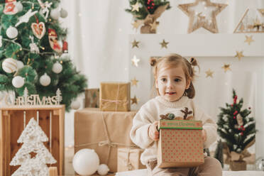 Nettes Mädchen sitzt mit Weihnachtsgeschenk in der Nähe von Dekoration zu Hause - EBBF08314