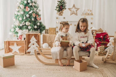 Glückliche Mädchen sitzen mit Geschenken in der Nähe von Weihnachtsdekoration zu Hause - EBBF08315