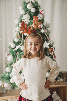 Happy girl standing with hands on hip in front of Christmas tree - EBBF08316