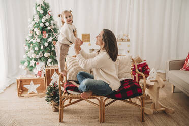 Lächelnde Mutter hält Hände und genießt mit Tochter auf Sessel zu Hause - EBBF08323