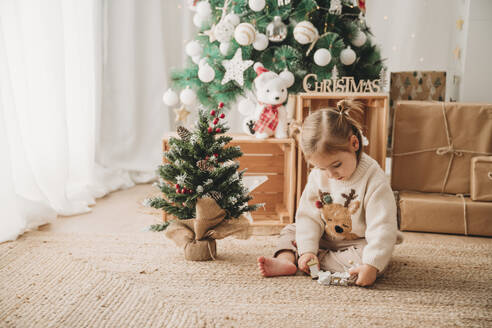 Mädchen spielt mit Weihnachtsschmuck in der Nähe von Baum zu Hause - EBBF08324