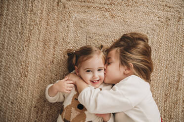 Girl kissing smiling sister lying on carpet at home - EBBF08337