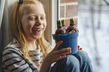 Glückliches Mädchen hält Hyazinthenzwiebeln und sitzt am Fenster - IHF01868