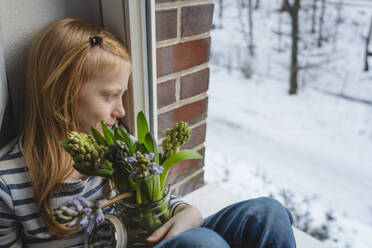 Mädchen hält einen Strauß Hyazinthenblüten in einem Glas und sitzt am Fenster - IHF01872