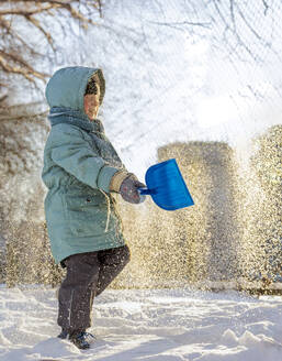 Junge trägt Winterjacke und wirft Schnee mit Spielzeugschaufel - MBLF00251