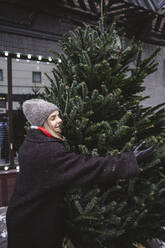 Smiling woman wearing knit hat and hugging Christmas tree - EYAF02947