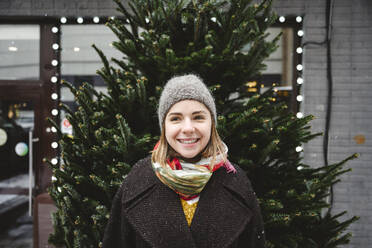 Happy young woman standing in front of Christmas tree - EYAF02948