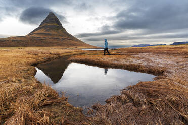 Frau geht vor dem berühmten Berg Kirkjufell spazieren, der sich im Wasser eines kleinen Sees spiegelt, Snaefellsnes-Halbinsel, Westisland, Island, Polarregionen - RHPLF31682