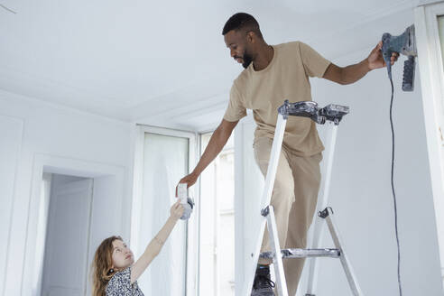 Woman giving ear muff to man standing on ladder and painting wall - DANF00028