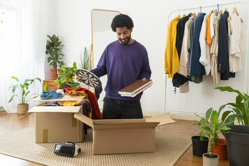 Smiling young man decluttering clothes and objects at home - VRAF00274