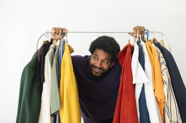 Happy young man looking through clothes rack at home - VRAF00280