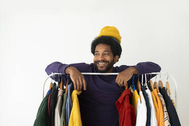 Smiling man wearing knit hat and leaning on clothes rack at home - VRAF00281