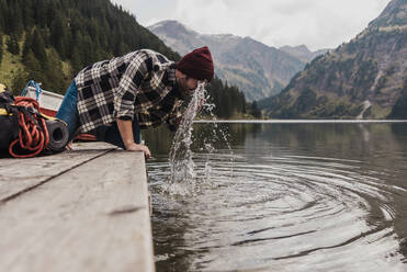 Junger Mann wäscht sich das Gesicht mit Seewasser vor einer Bergkulisse in Tirol, Österreich - UUF31096