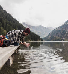 Glücklicher junger Mann, der das Wasser eines Sees in den Händen hält, in der Nähe der Berge in Tirol, Österreich - UUF31097