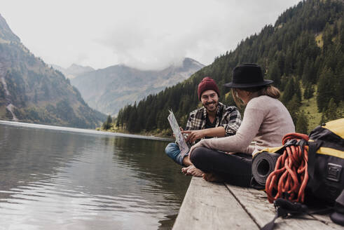 Glückliches junges Paar sitzt mit Landkarte auf einem Steg in der Nähe des Vilsalpsees und der Berge in Tirol, Österreich - UUF31098