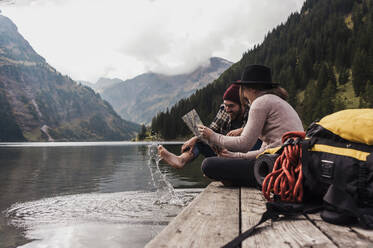 Glückliches Paar sitzt mit Landkarte auf einem Steg in der Nähe des Vilsalpsees und der Berge in Tirol, Österreich - UUF31099