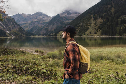 Thoughtful man with backpack standing in front of lake Vilsalpsee and mountains at Tyrol, Austria - UUF31101