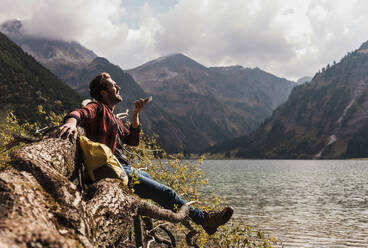 Junger Mann sendet eine Sprachnachricht über sein Smartphone auf einem umgestürzten Baumstamm in der Nähe des Vilsalpsees und der Berge in Tirol, Österreich - UUF31105