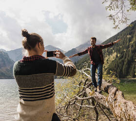 Junge Frau fotografiert einen glücklichen Mann, der auf einem umgestürzten Baumstamm in der Nähe des Vilsalpsees und der Berge in Tirol, Österreich, spazieren geht - UUF31108