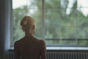 Nackte junge Frau mit blondem Haar schaut durch ein Fenster - AZF00599