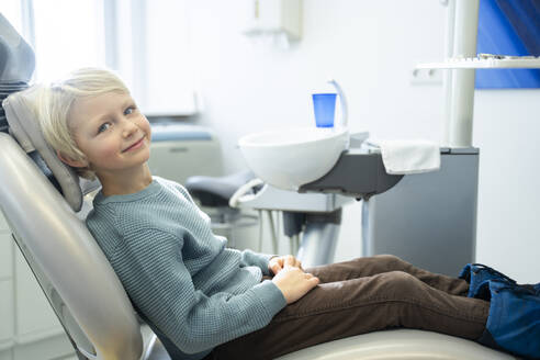 Lächelnder Junge sitzt auf einem Stuhl für eine zahnärztliche Untersuchung in einer Klinik - NJAF00750