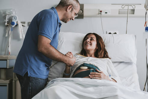 Happy man consoling pregnant woman on bed at hospital - EBBF08358