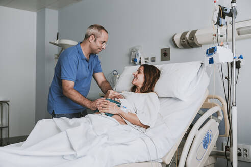 Smiling man consoling pregnant woman on bed at hospital - EBBF08359