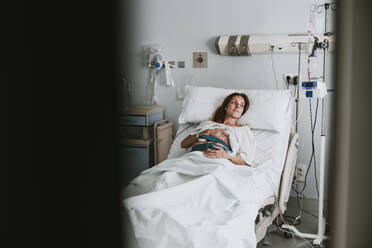 Smiling pregnant woman with hands on stomach lying on bed in hospital - EBBF08361