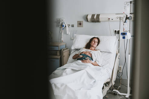 Smiling pregnant woman with hands on stomach lying on bed in hospital - EBBF08361