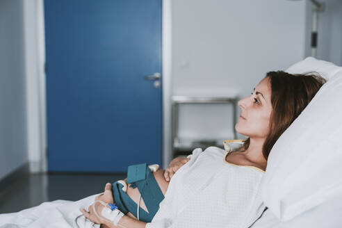 Thoughtful Pregnant woman with hands on stomach lying in hospital - EBBF08364