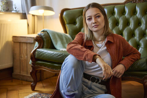 Smiling young woman sitting near sofa in living room at home - SUF00731