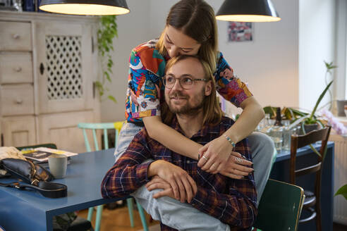 Young woman embracing man sitting on chair near dining table at home - SUF00735