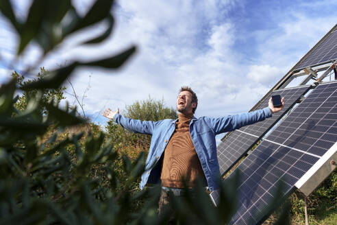 Mature man with arms outstretched screaming and standing near solar panels - JOSEF23110