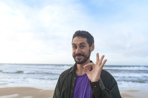 Smiling man gesturing OK sign in front of sea - JOSEF23132