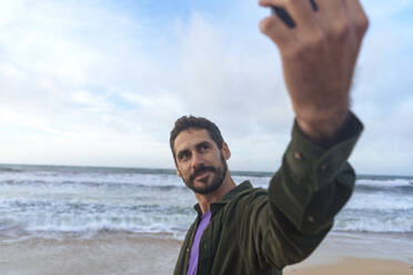 Man taking selfie through smart phone in front of sea at beach - JOSEF23135