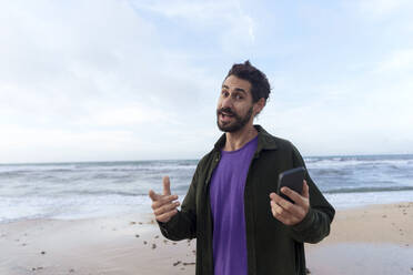 Glücklicher Mann mit Gesten und Smartphone am Strand - JOSEF23142